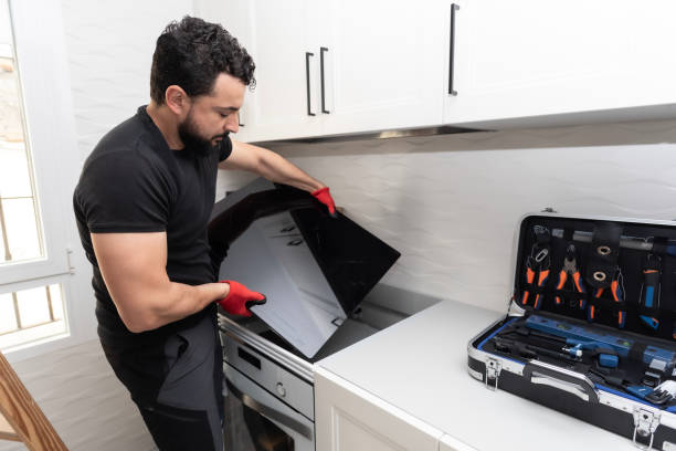 A man repairs a laptop on a kitchen counter, surrounded by tools and appliances related to range and cooktop installation.