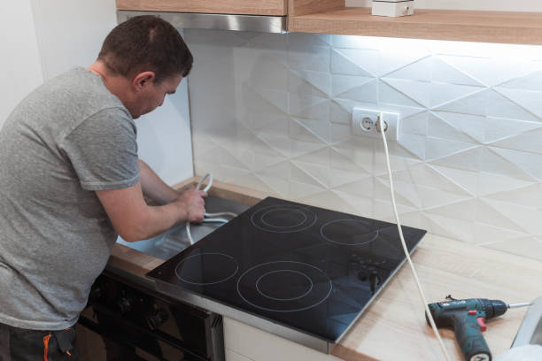 In a kitchen, a man is focused on fixing a laptop, with installation tools for a range and cooktop nearby.
