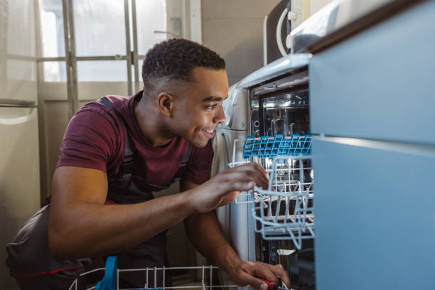 A man is install a dishwasher, focused on connecting the plumbing and electrical components.