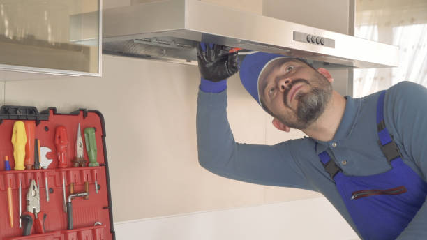 A man in blue overalls is engaged in fixing a Range Hood Installation, working on a kitchen appliance in a bright kitchen environment.