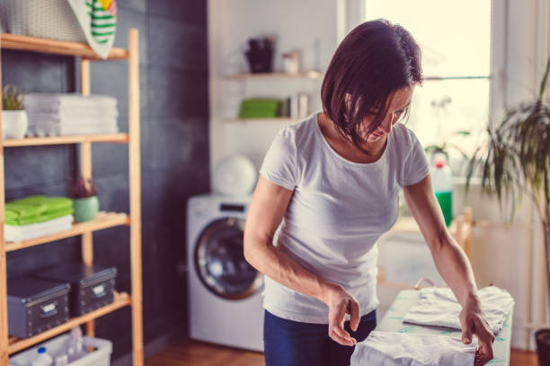 A woman is folding freshly laundered clothes in a tidy laundry room with a washer and dryer set in the background.
