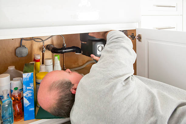  A man repairs a water heater beneath a sink, focusing on the installation of a garbage disposal unit.
