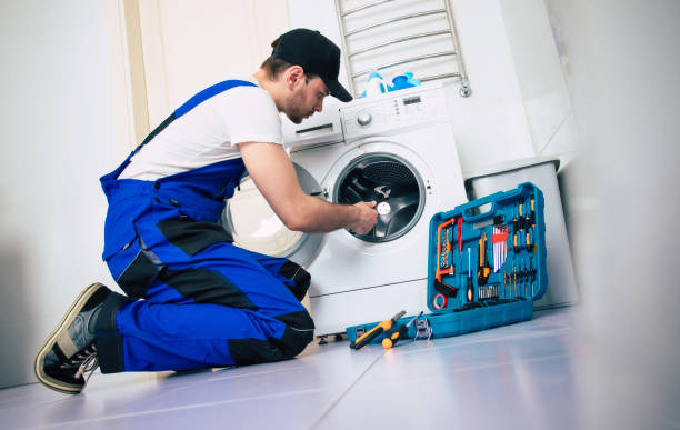 A man wearing overalls works on a washing machine indoors, engaged in Indoor Appliance Installation and repair tasks.