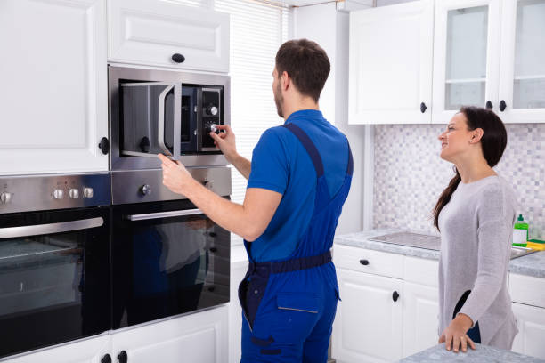 A man and woman in blue overalls stand in front of an over the range microwave, discussing its features.