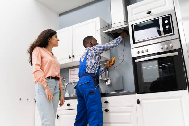 Two individuals in blue overalls stand before an over-the-range microwave, engaged in conversation about its use.