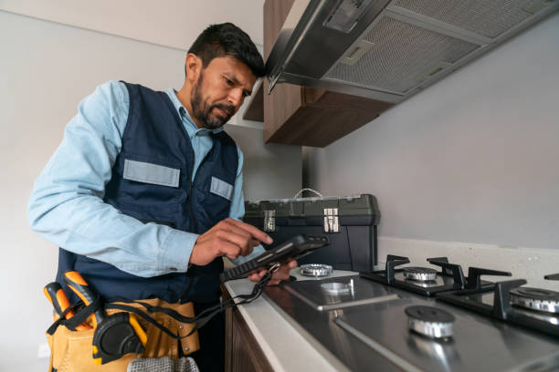 A man in a vest and hard hat is using a cell phone while overseeing appliance installation work.