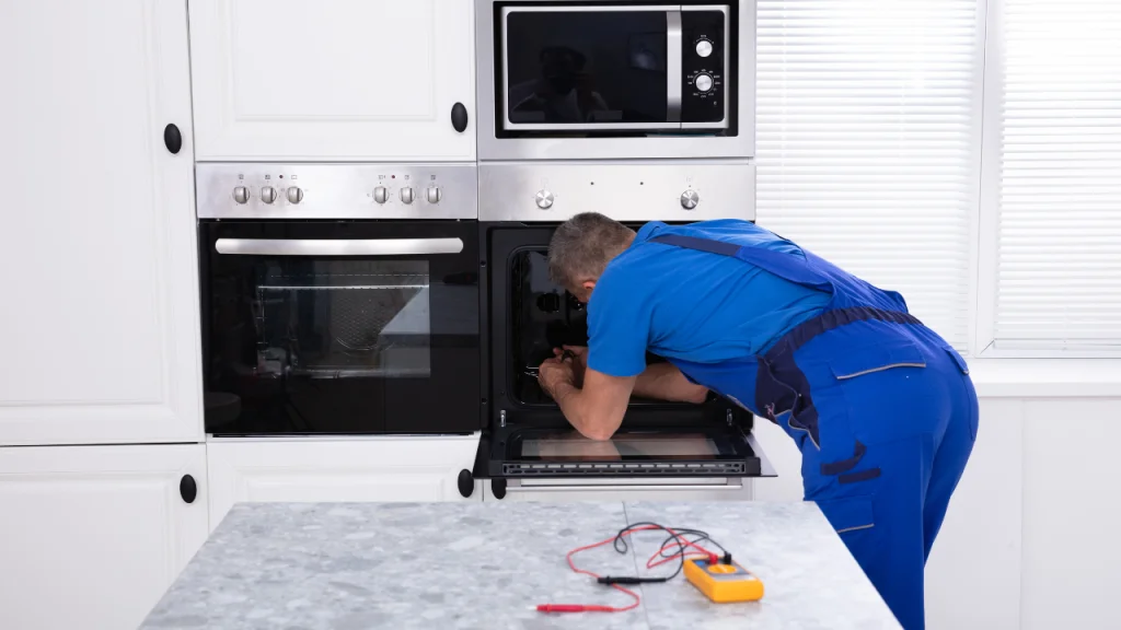 A technician in blue overalls leans into an open wall oven to perform repairs, with a digital multimeter resting on the counter in the foreground.
